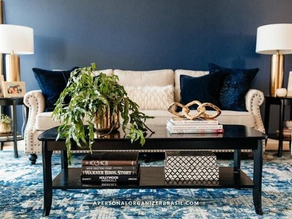 A stylish living room shows how to como organizar a sala de estar, with a beige sofa, navy blue pillows, and a dark coffee table topped with a potted plant, decorative items, and books. Two table lamps frame the deep blue accent wall.