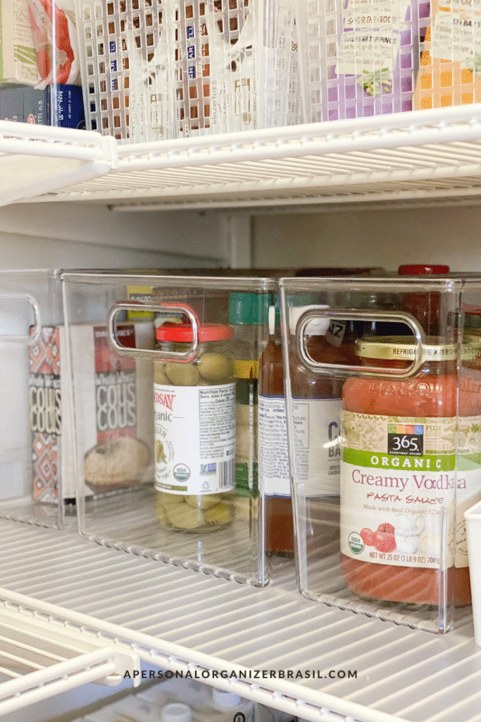 Clear plastic storage bins on white wire shelves hold jars, bottles, couscous, and organic pasta sauce for a neatly organizar a despensa. The website "apersonalorganizerbrasil.com" appears at the bottom.