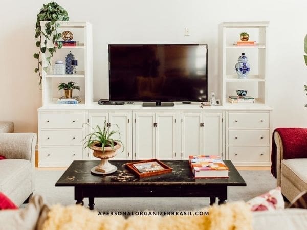 A sala organizada conta com uma TV sobre um aparador branco, ladeada por prateleiras com plantas, livros e vasos. Uma mesa de centro organizada exibe livros e uma planta diante de um sofá bege com manta vermelha, criando um ambiente acolhedor.