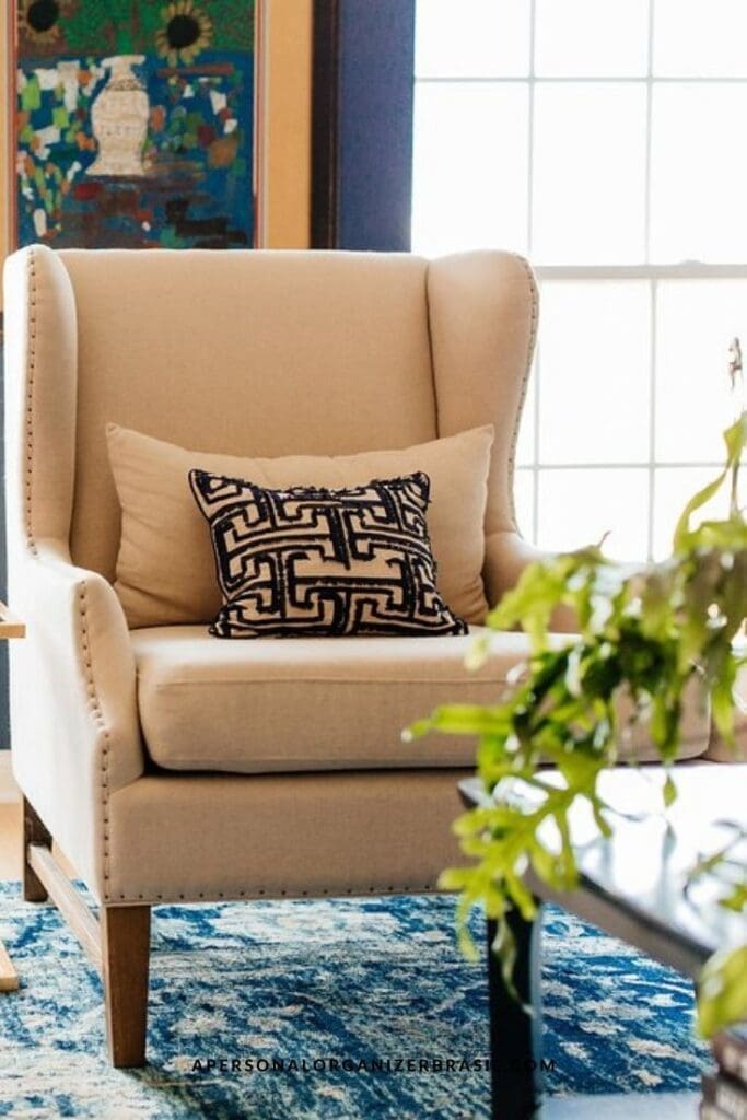 Como Organizar a Sala de Estar. A beige wingback armchair with wooden legs and stud detailing sits on a blue patterned rug—perfect inspiration for quem quer aprender como organizar a sala de estar. Two patterned cushions and a leafy green plant complete the inviting scene.