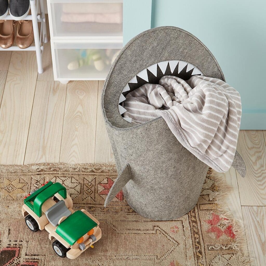 A gray shark-shaped laundry basket helps with organizing, holding a striped blanket on a rug with wooden floors. Next to it is a green and wood-colored toy truck, while shoes and storage bins in the background aid in tidying the space.