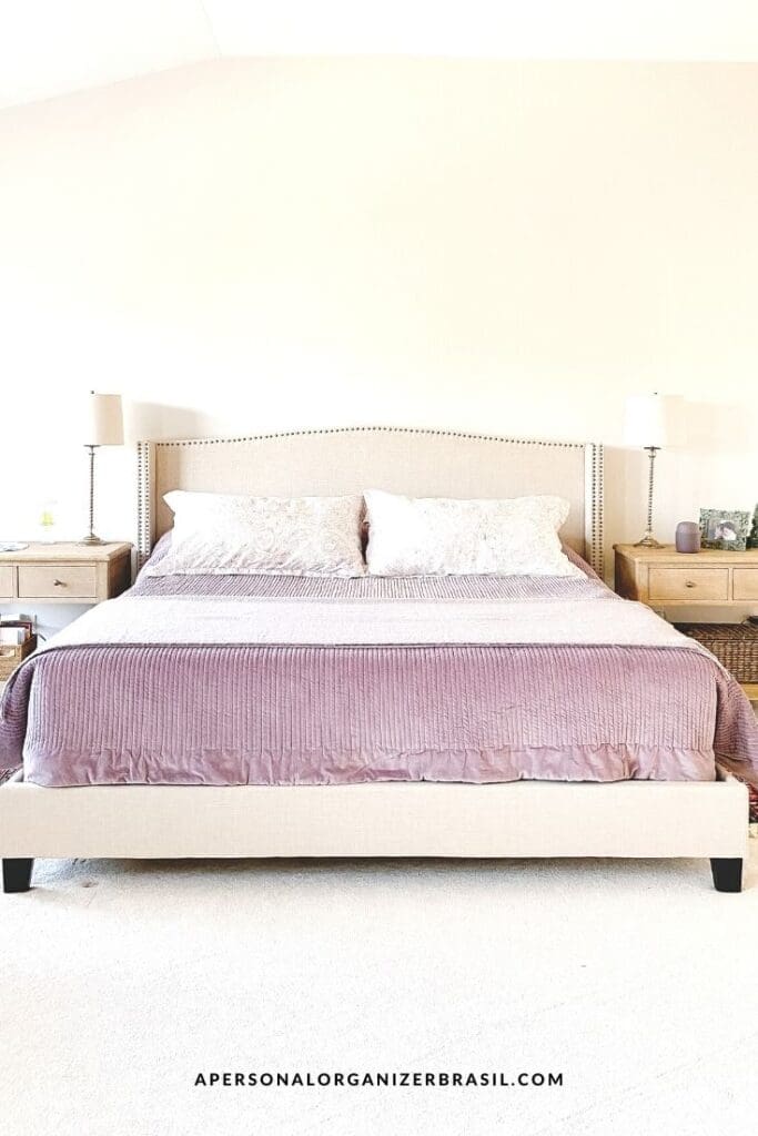 A neatly made bed with a beige upholstered headboard, lavender bedding, and two matching pillows. Two wooden nightstands with lamps and decor items add charm to this bright, cozy room—perfect for those who love organizing and a clutter-free space.