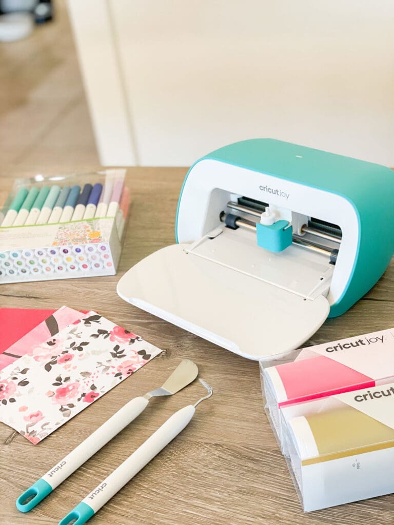 A Cricut Joy cutting machine sits on a wooden table, surrounded by colorful pens, patterned paper, vinyl rolls, and organizing tools—everything you need for a perfectly organized crafting project.