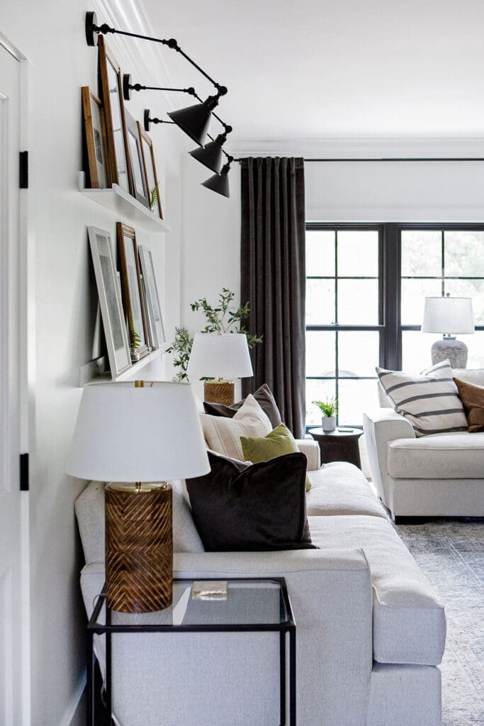 A modern living room with light-colored sofas, black-framed windows, wall-mounted lamps, framed art on picture ledges, side tables with lamps, and neutral-toned decor with green and brown accent pillows.