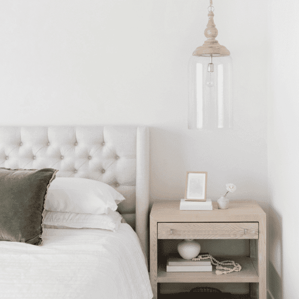 A minimalist bedroom features a gray tufted headboard and a neatly made bed with two pillows. A wooden nightstand, perfect for organizing products, holds a small framed picture, a decorative object, and a hanging glass pendant light above it against the white wall.
