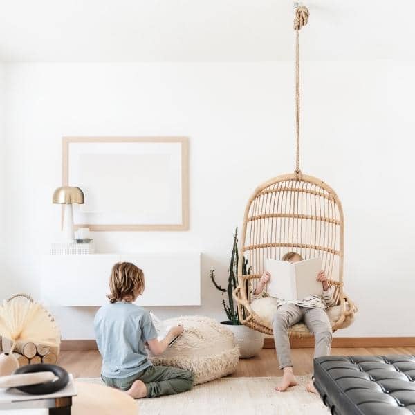 In a minimalist living room, two children immerse themselves in books—one seated on the floor and the other swaying gently in a hanging wicker chair. Surrounded by sleek organizing products, cacti, and modern decor in neutral tones, they enjoy their serene reading haven.