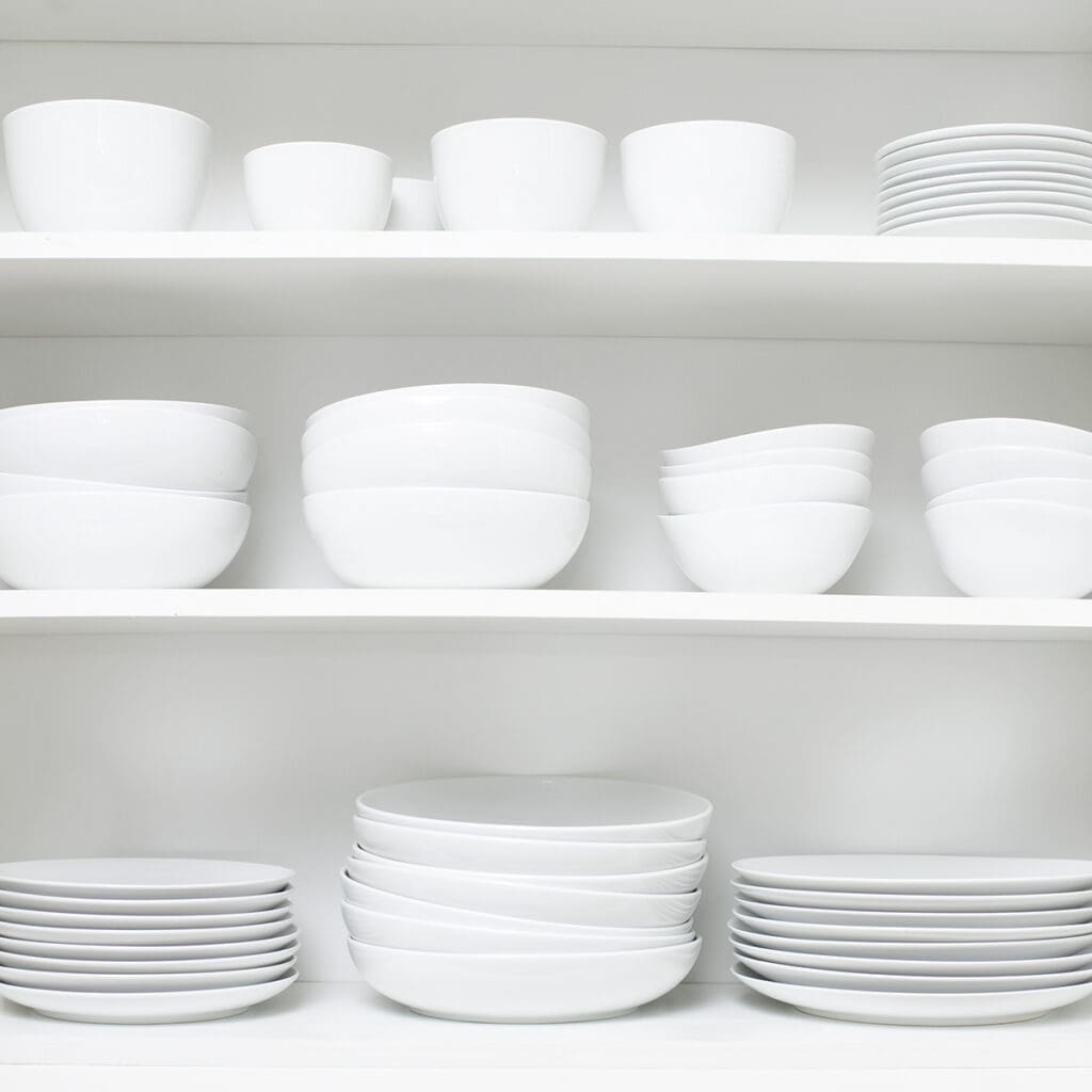 Shelves filled with neatly stacked white ceramic dishes, including plates, bowls, and mugs, showcase expert organizing. The minimalist arrangement reflects an organized and uncluttered kitchen space.