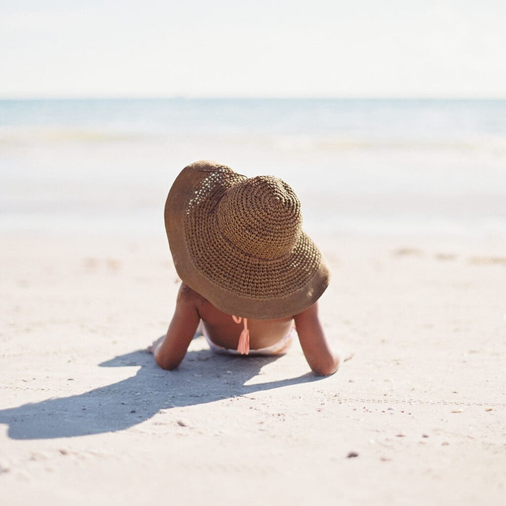 A person lies on a sandy beach facing the ocean, wearing a large straw hat, as the sun entertains with its gentle warmth. Shadows dance while the calm sea and clear blue sky decorate the scene with serene beauty.