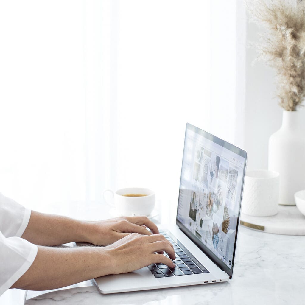 A person is typing on a laptop at a bright, white desk with coffee, a vase with dried flowers, and decorative objects—perhaps searching for a curso personal organizer as various images appear on the screen.