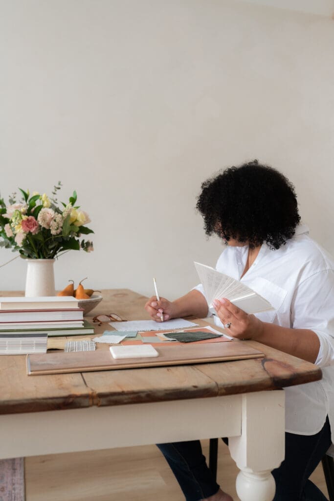 A person with curly hair sits at a wooden table, jotting down notes for their curso personal organizer with a white pen. Surrounded by stacks of books, notes, and a vase of flowers, they wear a white shirt and focus intently on perfecting their work.