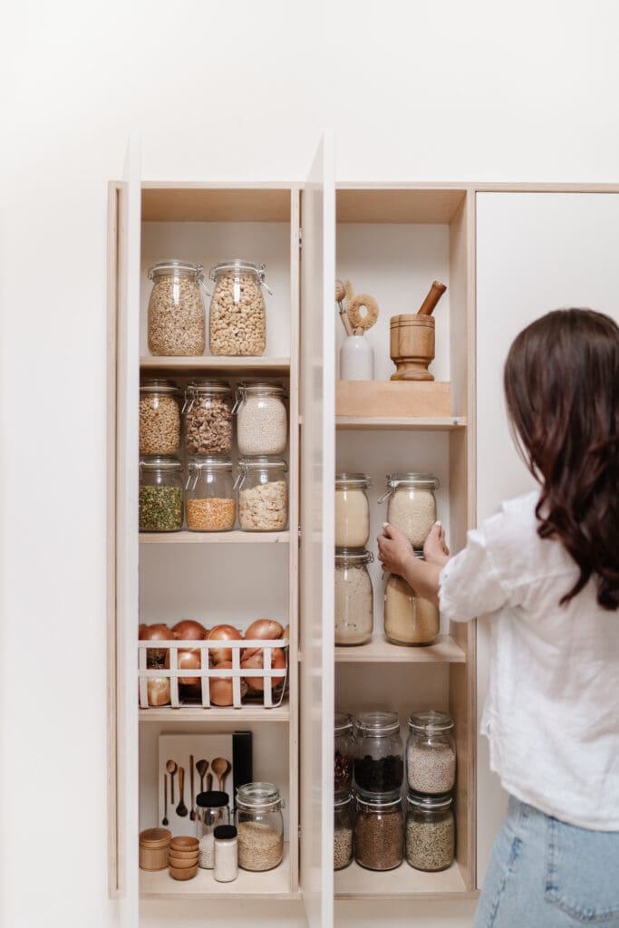 A person is organizing products on a pantry shelf, arranging glass jars filled with grains and spices. The shelves display legumes, spices, and a basket of onions. Wooden utensils and a mortar and pestle add to the kitchen's charm.