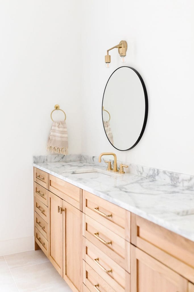 A bathroom vanity with light wood cabinets, gold hardware, a marble countertop, gold faucet, round mirror, gold wall sconce, and a striped hand towel hanging on a gold ring against a white wall.