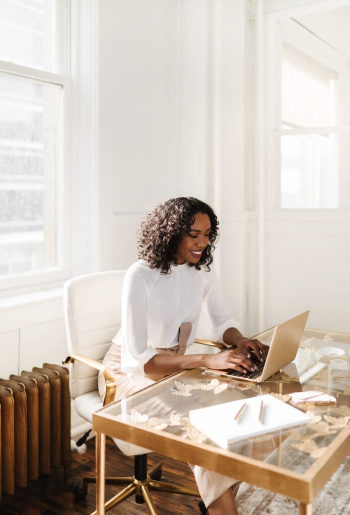 A person with curly hair, wearing a white blouse, sits at a desk using a laptop. The room is bright with natural light from large windows, and there is a radiator and a white chair. The desk holds decorative items befitting an organized personal organizer.