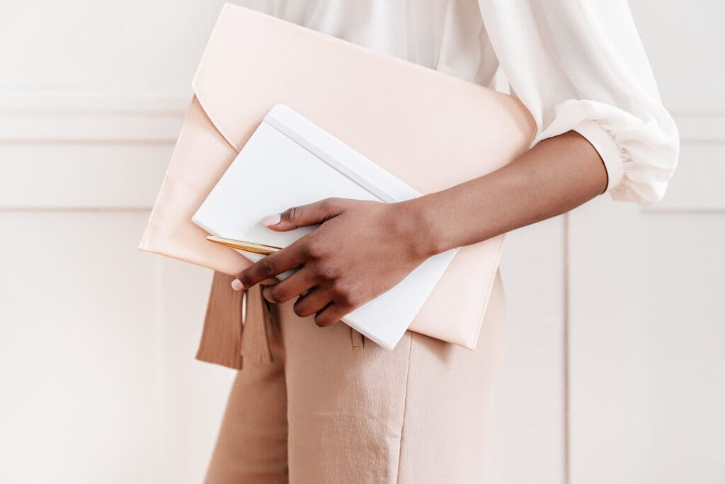 Person holding a white notebook and a beige envelope-style clutch, possibly containing formulários e modelo de contrato para personal organizer. They are wearing light brown pants and a white blouse against a softly lit, neutral-toned wall.