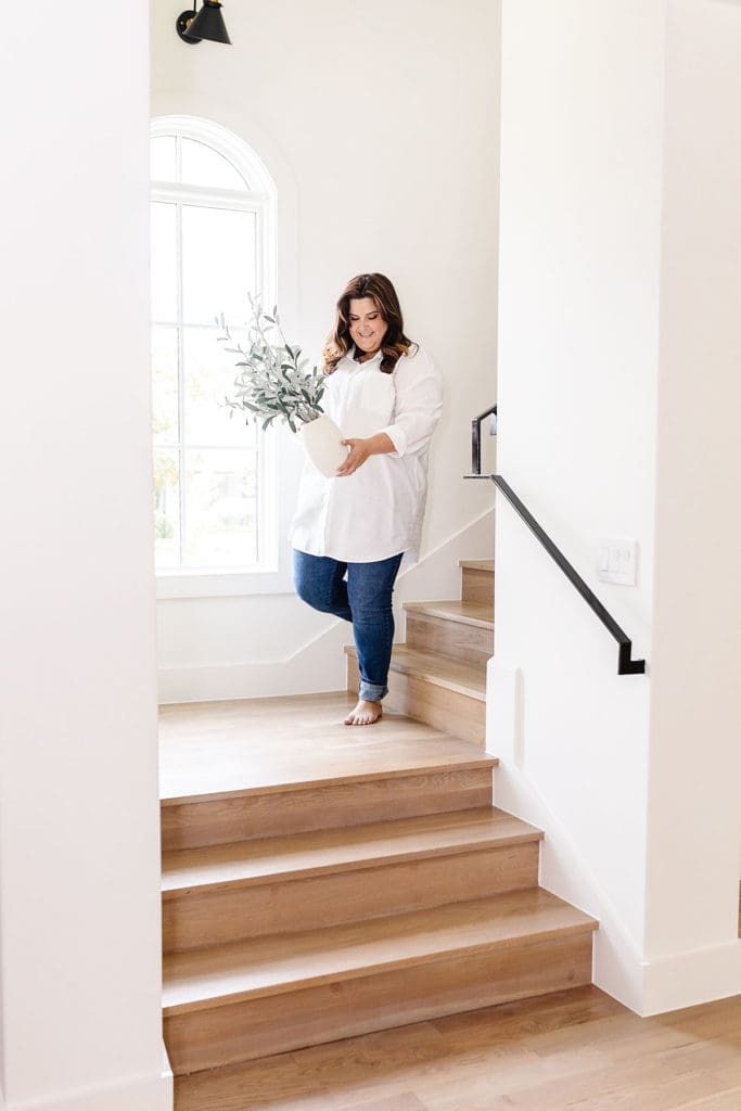 A woman in a white shirt and blue jeans walks down wooden stairs in a bright, modern casa organizada, holding a white vase with greenery. A large window lets in natural light behind her.