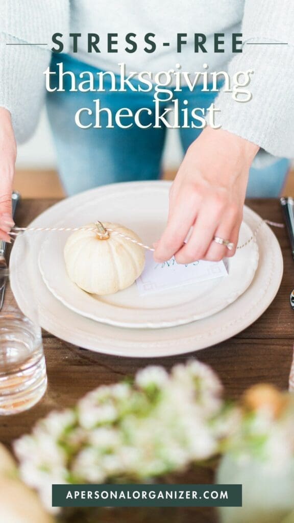 Close-up of a Thanksgiving table setting with a white pumpkin on a plate, tied with twine holding a note. A person's hands are adjusting it. Text on image reads "Stress-Free Thanksgiving Checklist" and "apersonalorganizer.com" at the bottom.
