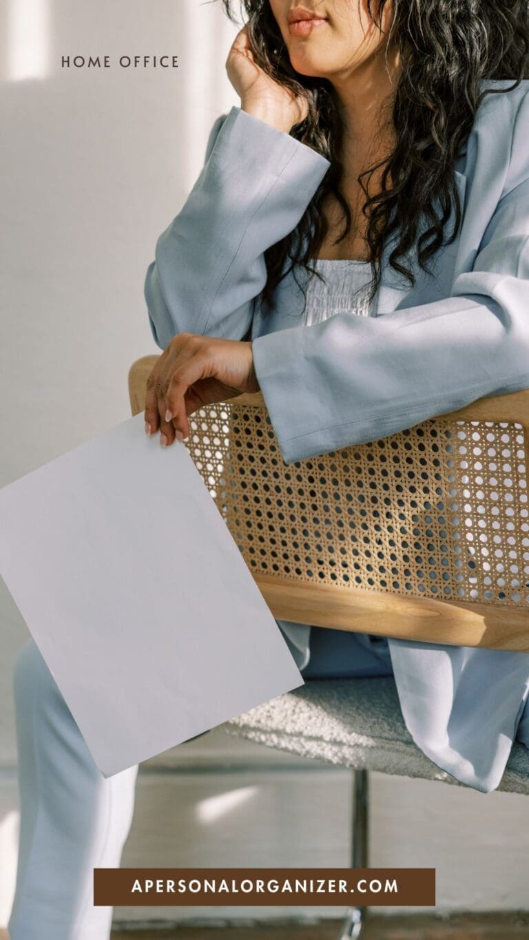 A woman in a light blue suit sits on a woven chair, holding a blank sheet of paper—perhaps considering an important document organizer or filing system. Sunlight casts shadows on her and the chair. Text reads "HOME OFFICE" and "APERSONALORGANIZER.COM.