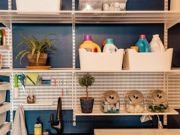 A laundry room with white shelves holding detergent bottles in white bins, two potted plants, and glass containers with laundry pods and clothespins offers inspiration beyond washing—much like tips on como organizar a sala de estar, it brings order and style to daily routines.