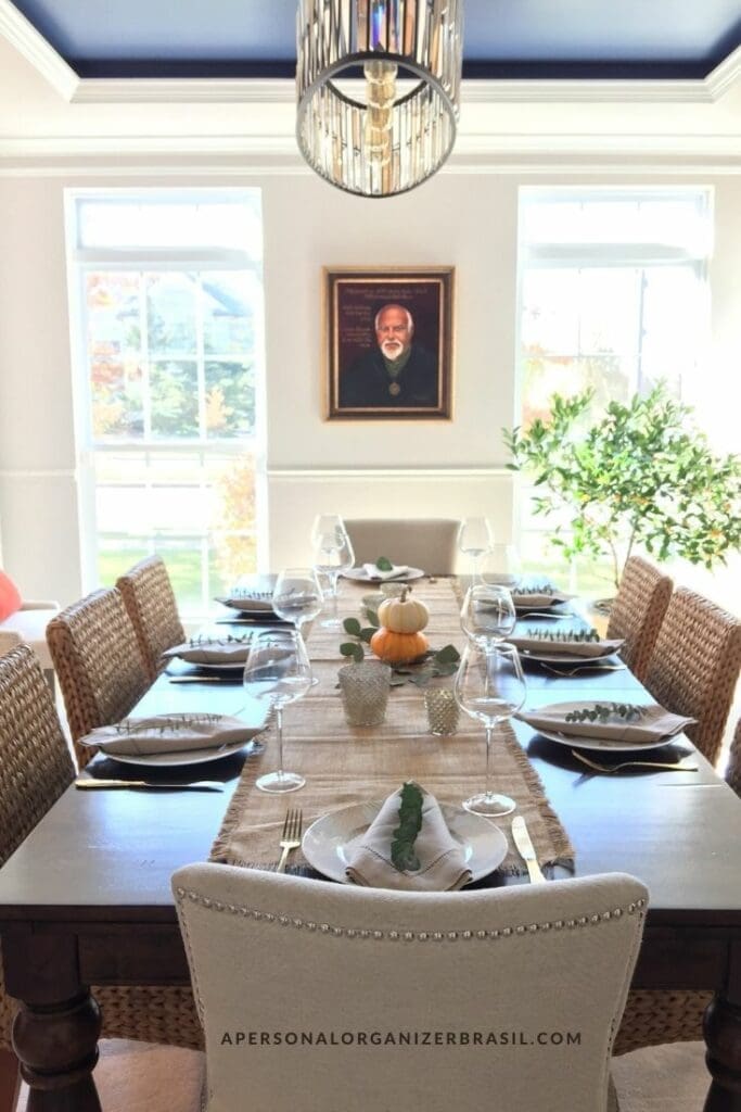 Bright sala de jantar with a long table set for eight, decorated with a small white pumpkin centerpiece and wine glasses. Woven chairs surround the table, and a portrait hangs on the wall in front of large windows.