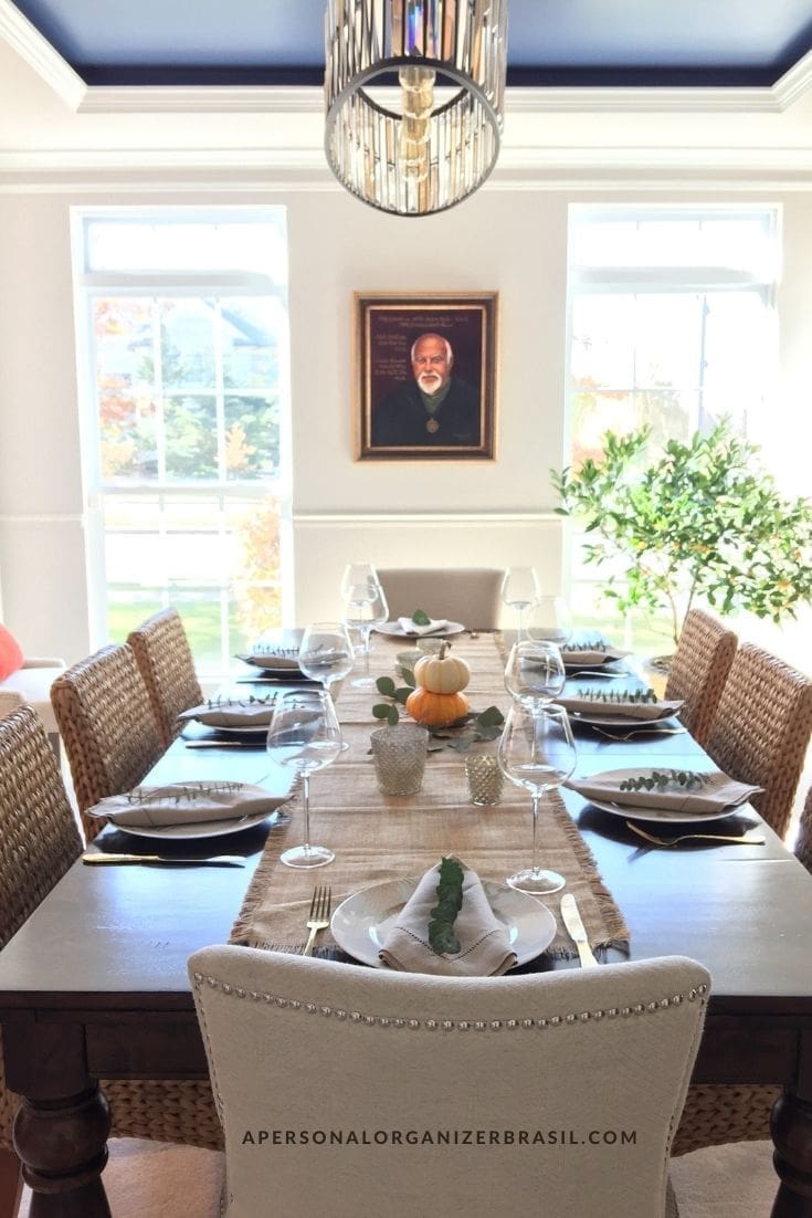 Bright sala de jantar with a long table set for eight, decorated with a small white pumpkin centerpiece and wine glasses. Woven chairs surround the table, and a portrait hangs on the wall in front of large windows.