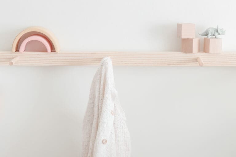 A light wooden wall peg rail with a white textured cloth hanging, pastel wooden stacking arches, wooden blocks, and a small toy dinosaur on top creates a charming cenário para o quarto das crianças against a plain white background.