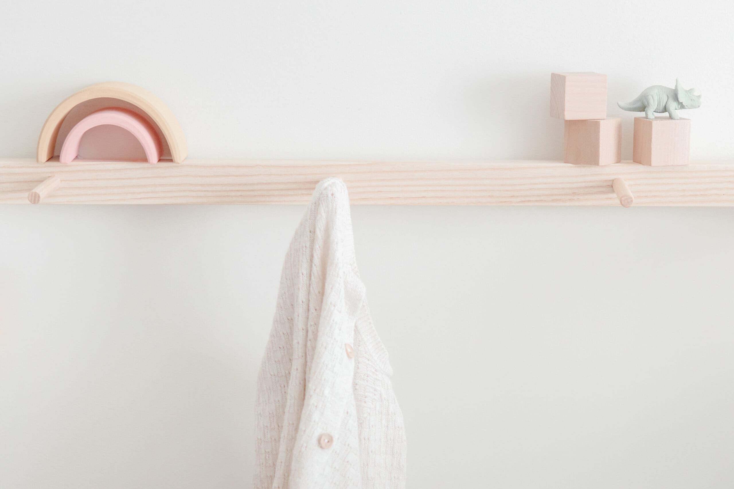 A light wooden wall peg rail with a white textured cloth hanging, pastel wooden stacking arches, wooden blocks, and a small toy dinosaur on top creates a charming cenário para o quarto das crianças against a plain white background.