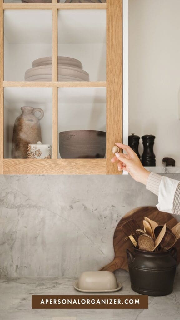 A hand opens a wooden kitchen cabinet with stacked ceramic dishes inside—perfect for decluttering for a move. On the marble counter below are wooden utensils in a container, a butter dish, and cutting boards. Text at the bottom reads "apersonalorganizer.com".
