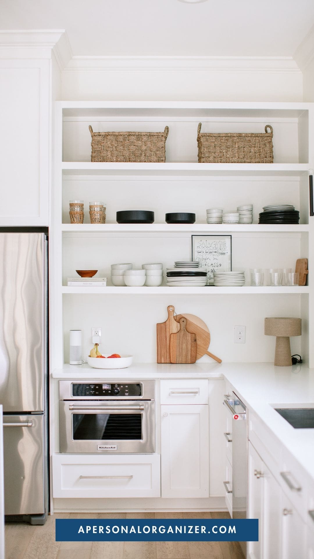 A modern white kitchen with open shelves displaying dishes, bowls, cups, and two woven baskets; built-in microwave, wooden cutting boards, and a lamp on the counter next to the refrigerator highlight thoughtful organization.