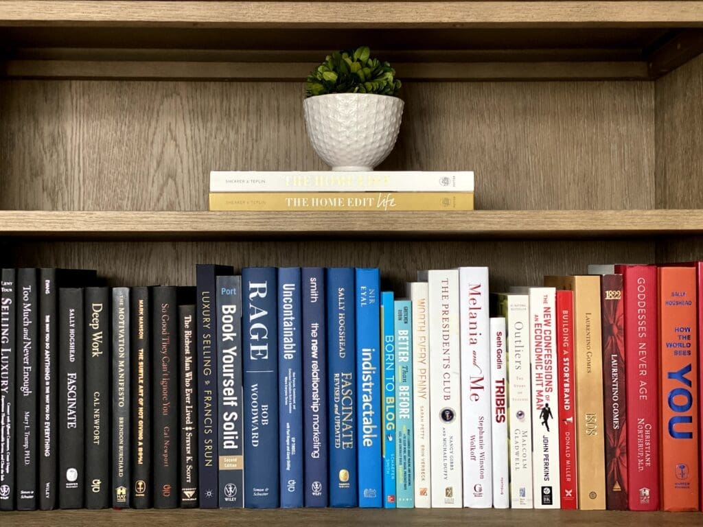 Books of various colors are neatly arranged on two wooden shelves, adding charm to the home office. On the top shelf, two light-colored books are stacked horizontally with a small green potted plant on top. The lower shelf displays a row of upright books.