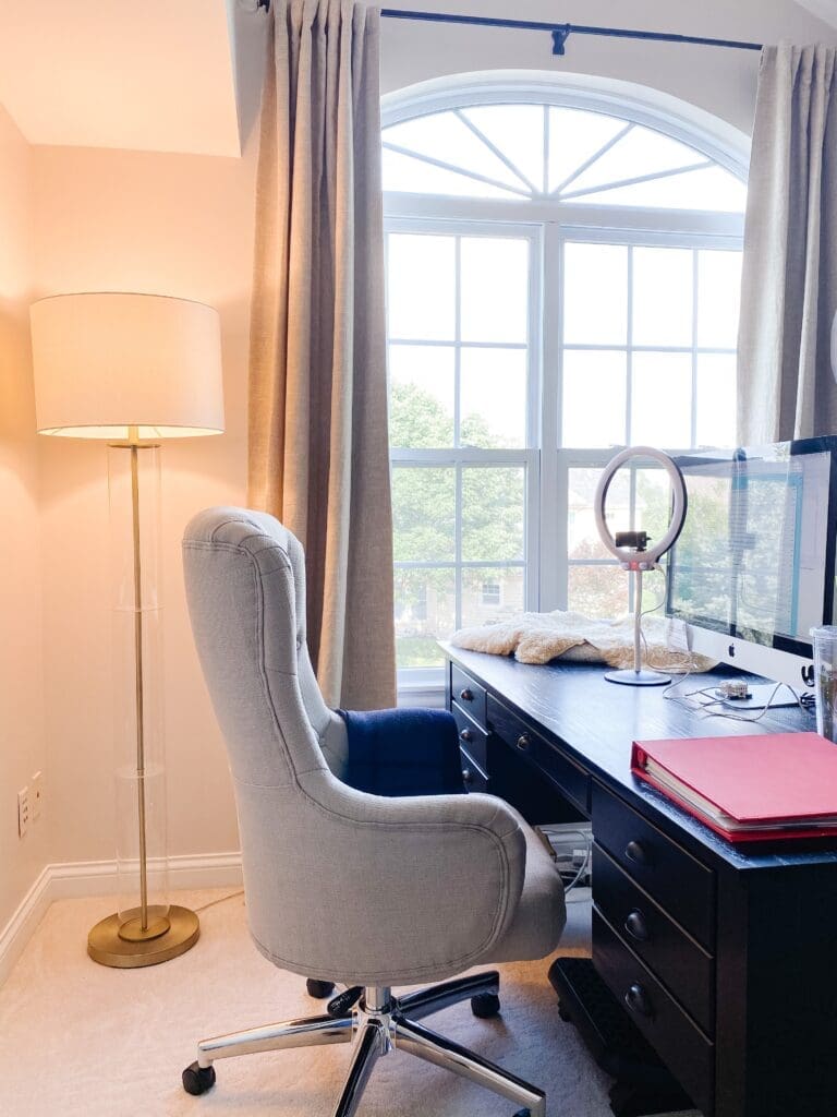 A bright home office with a tall window, an upholstered swivel chair at a dark desk, a floor lamp, computer monitor, ring light, mug, red folder, and beige curtains. Trees are visible outside this inviting home office space.