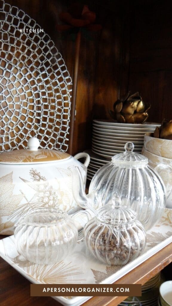 A close-up of a shelf with decorative plates, a teapot, matching cups, and glass jars on a white tray, all featuring elegant gold and white designs. The text reads "KITCHEN" and "APERSONALORGANIZER.COM".