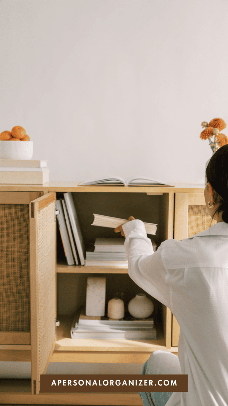 A person in a white shirt organizes books on a wooden cabinet shelf, creating an organized home. The cabinet top holds a bowl of oranges and a vase with orange flowers. Text at the bottom reads “APERSONALORGANIZER.COM.”.