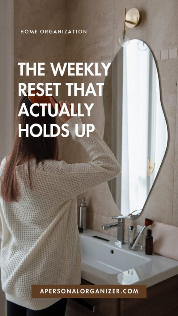 Woman in a cream sweater stands in a modern bathroom, adjusting her hair in the mirror. Text on image reads: “The Weekly Reset review: Home Organization That Actually Holds Up.”.