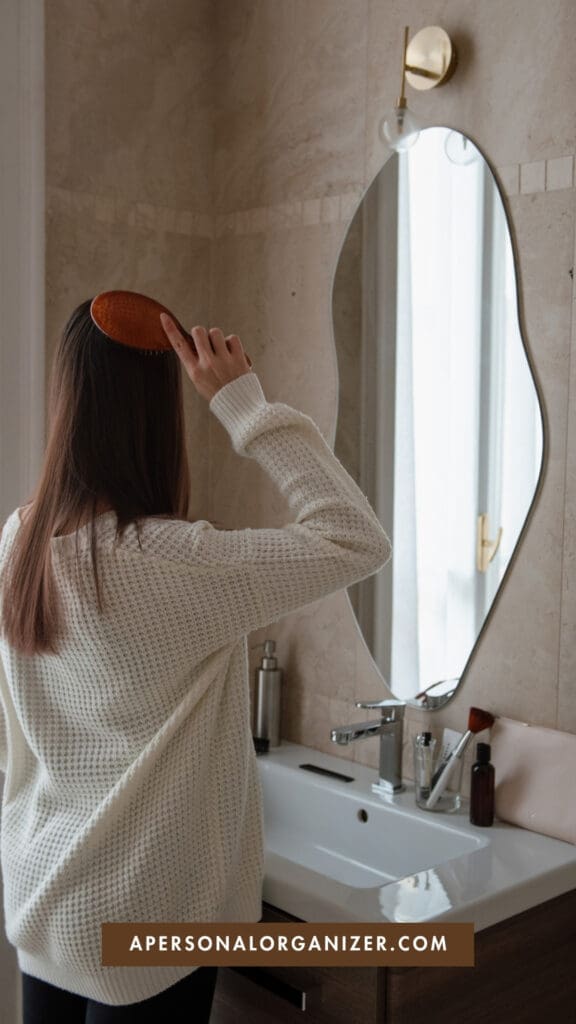A person with long hair, wearing a white sweater, brushes their hair in front of a bathroom mirror as part of their Weekly Reset routine. The bathroom has a sink, toiletries, beige tiled walls. “APERSONALORGANIZER.COM.”.