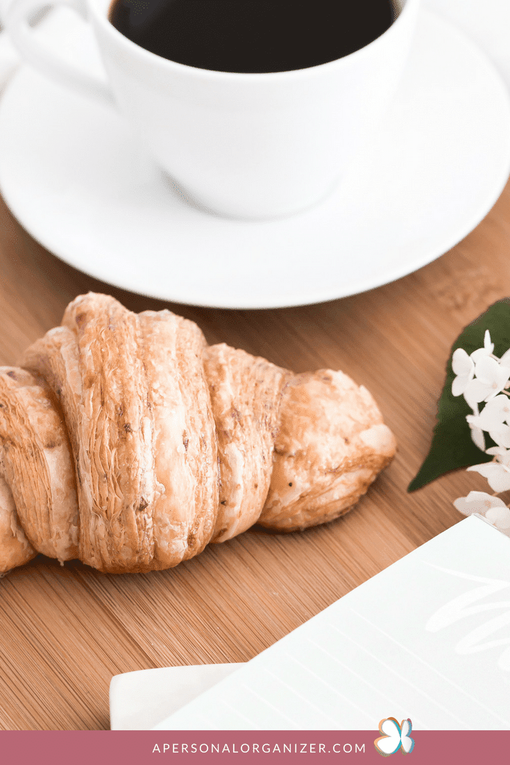 A croissant sits on a wooden table next to a white cup and saucer filled with coffee. Beside the croissant, you'll find a white notebook and some small white flowers—perfect elements for organizing morning routines. A website link, "apersonalorganizer.com," is visible at the bottom of the image.