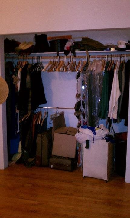 A cluttered closet with a wooden floor awaits organizing. Clothes hang on two racks, surrounded by boxes and bags piled at the bottom. Some garments are preserved in clear plastic, while a straw hat hangs casually on the left side.