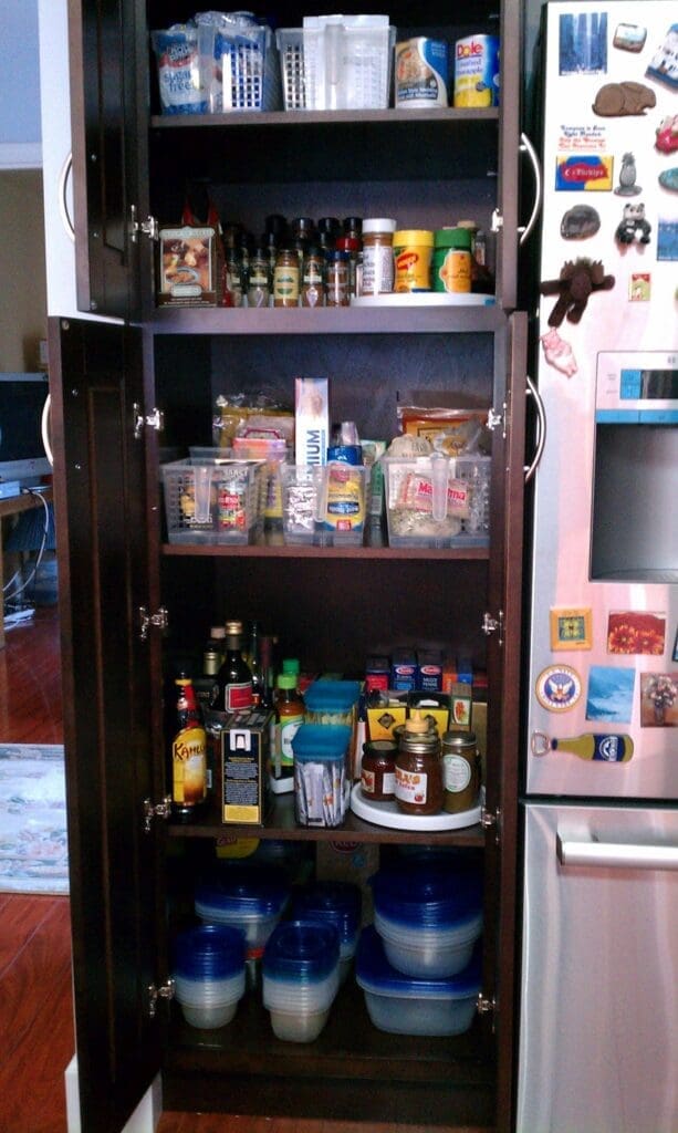 A tall pantry with open dark wood doors reveals impeccably organized shelves filled with various food items, spices, canned goods, plastic containers, and sauces. The fridge beside it is a canvas of magnets and stickers, capturing a lively contrast to the tidy organizing within.