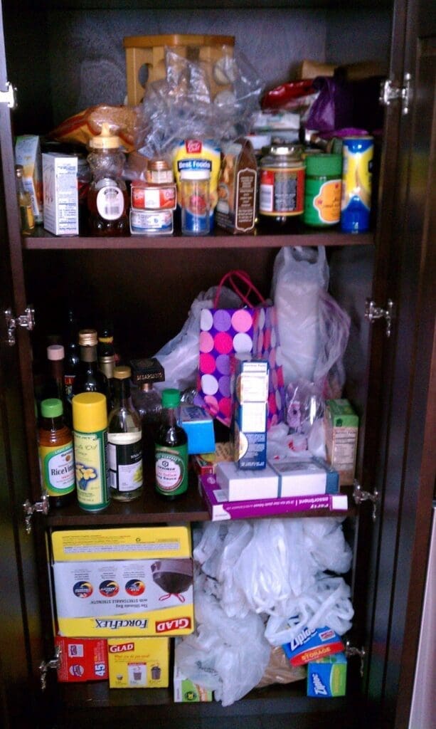 A cluttered pantry with various items, including jars, cans, boxes, and bags. Amidst the chaos, an organizing miracle takes shape as neatly arranged shelves boast bottles of oil and packaged goods. A colorful shopping bag adds a vibrant touch to this transformed space.
