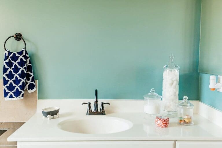 A tidy bathroom showcases a white countertop and sink, perfect for bathroom organizing. A black faucet is centered above the sink. To the right, glass jars hold cotton balls and essentials. A blue and white patterned towel elegantly hangs on a ring to the left on the green wall.