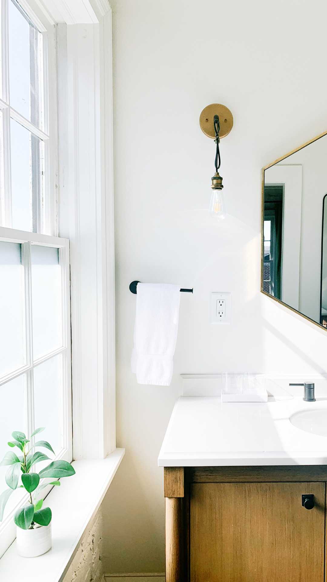 Organized bathroom with a large window on the left, a plant on the windowsill, a white towel neatly hanging on a black rod, and a square mirror above a wooden vanity with a white countertop. A bulb with a vintage fixture is mounted on the wall.