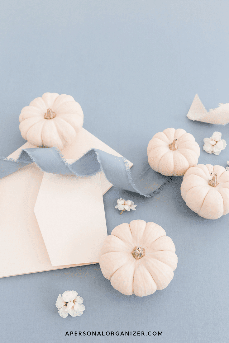 A flat lay of small white pumpkins arranged on a light blue background with pale pink envelopes and a light blue fabric ribbon invites you to give thanks. White flowers are scattered among the pumpkins and envelopes. The website "apersonalorganizer.com" is displayed at the bottom.