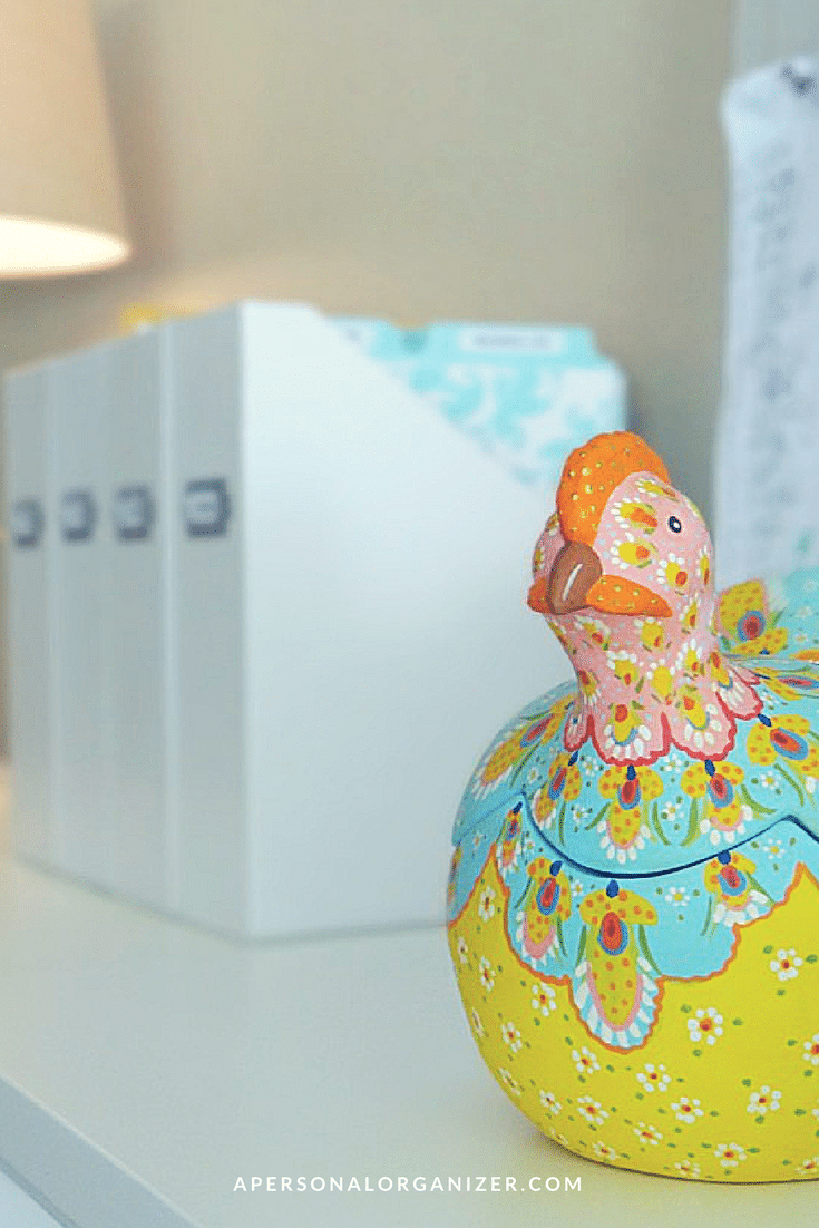 A decorative ceramic container shaped like a colorful bird sits on a white desk. Behind it are organized white file holders with labels, perfect for home office organization ideas. The background includes a lamp and a soft-focus light blue patterned cloth. The text "APERSONALORGANIZER.COM" is visible at the bottom.