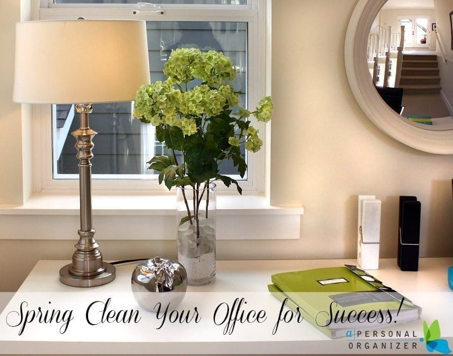 A well-organized white desk with a silver lamp, a clear vase of green hydrangeas, a stack of green folders, and office supplies. A round mirror and window are visible in the background. Text at the bottom reads: "Spring Cleaning Your Office for Success!