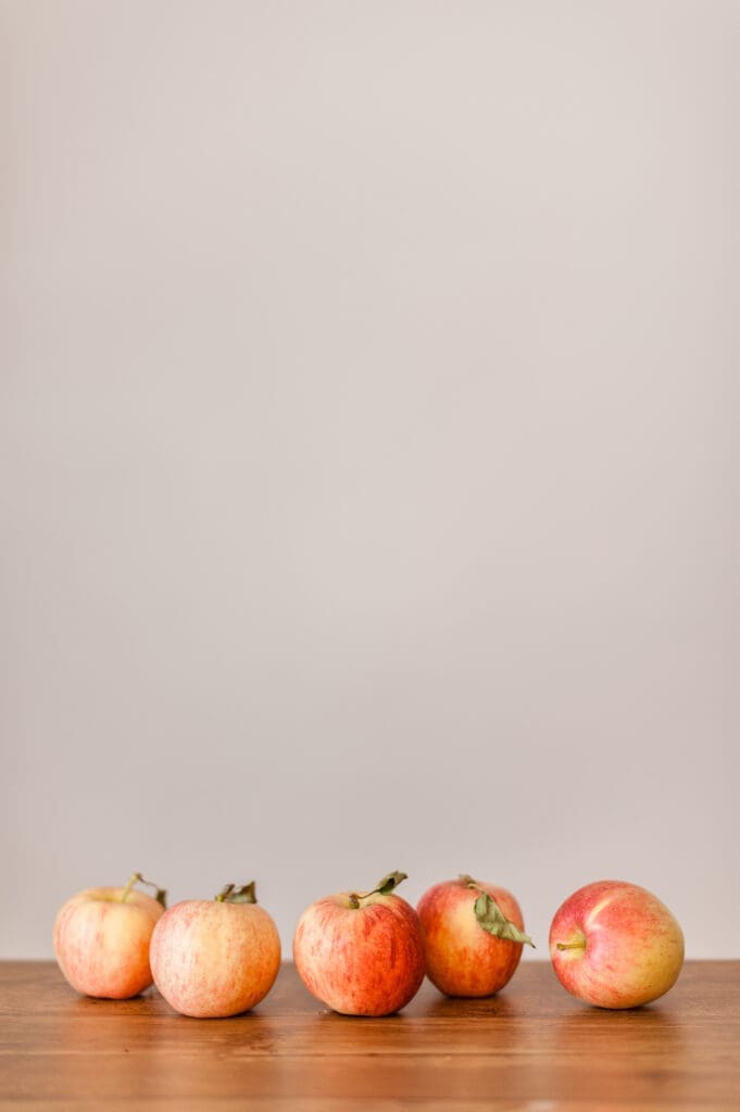 Five red and yellow apples are arranged in a row on a wooden surface, resembling the orderly neatness of school papers organization, set against a plain, light-colored background.