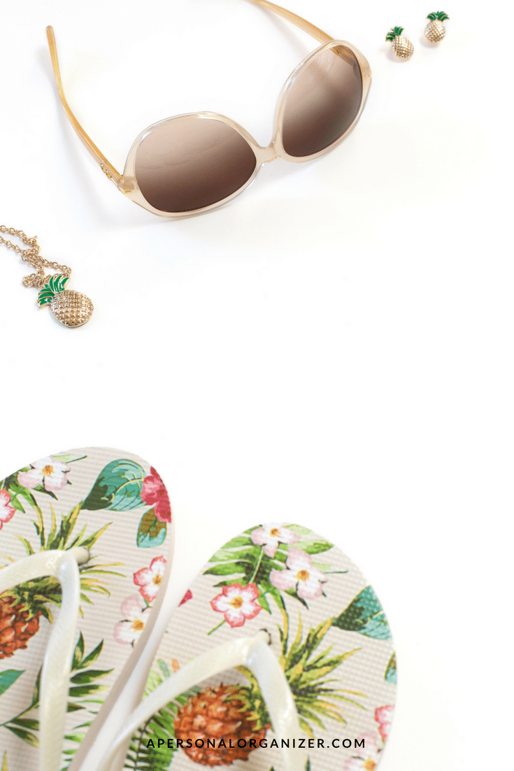 A pair of large brown-lensed sunglasses, gold pineapple-themed earrings, a matching necklace, and floral flip-flops with pineapples and flowers, all arranged on a white background. Complete your vacation checklist at "apersonalorganizer.com," featured at the bottom.