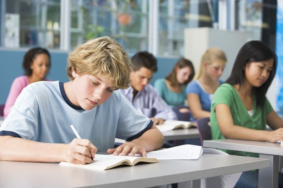 A classroom with several students sitting at desks, focused on writing in their notebooks. The nearest student, a boy with blond hair in a light blue shirt, writes in an open book with a pen. Others in the background are also engaged in their work, possibly jotting down time management tips.