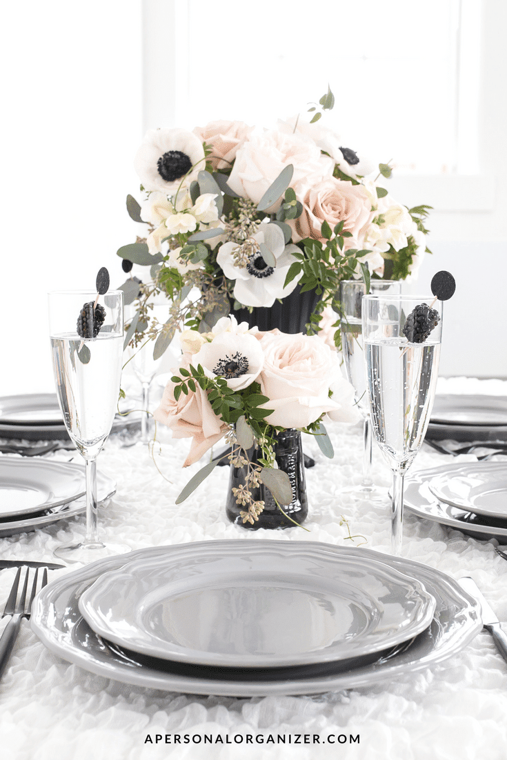 Elegant fall table decorations feature a centerpiece of white and blush roses with greenery. Four place settings with grey plates and black cutlery surround the arrangement. Each place includes a champagne flute with a small black decorative detail inside, adding a touch of autumnal charm.