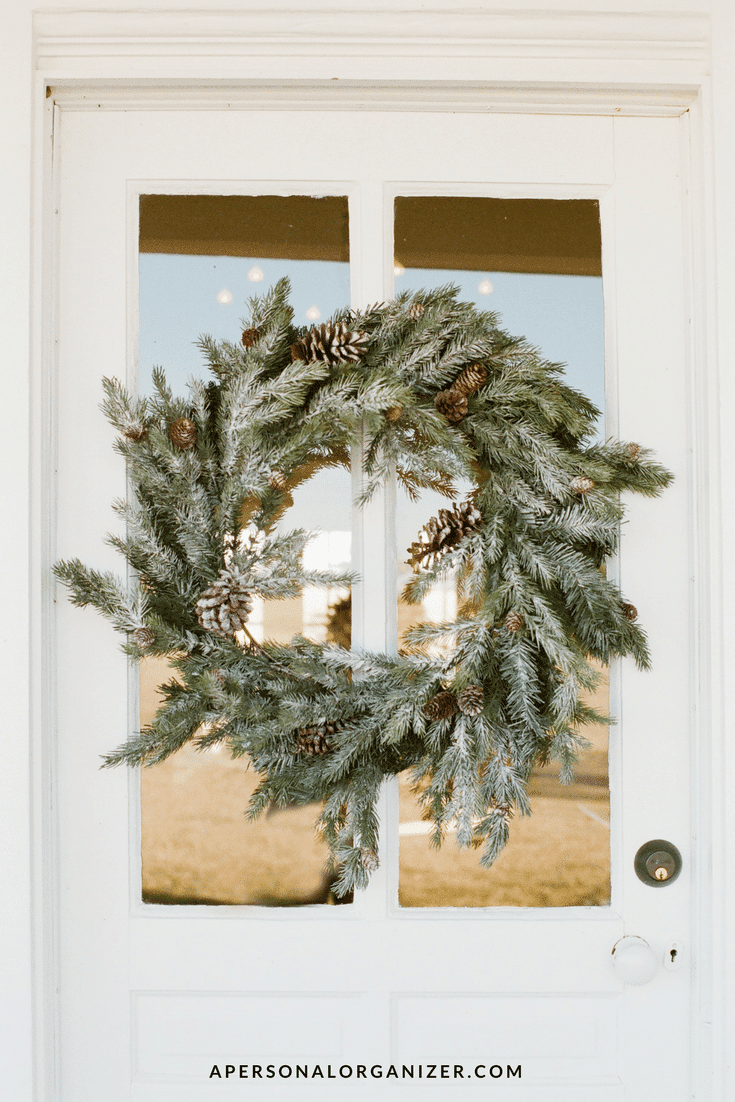 A large green holiday wreath with pinecones, dusted with snowy white, hangs on a glass window of a white door. Perfect for getting your home ready for the holidays, the wreath is centered, and the website "apersonalorganizer.com" is visible in small text at the bottom.