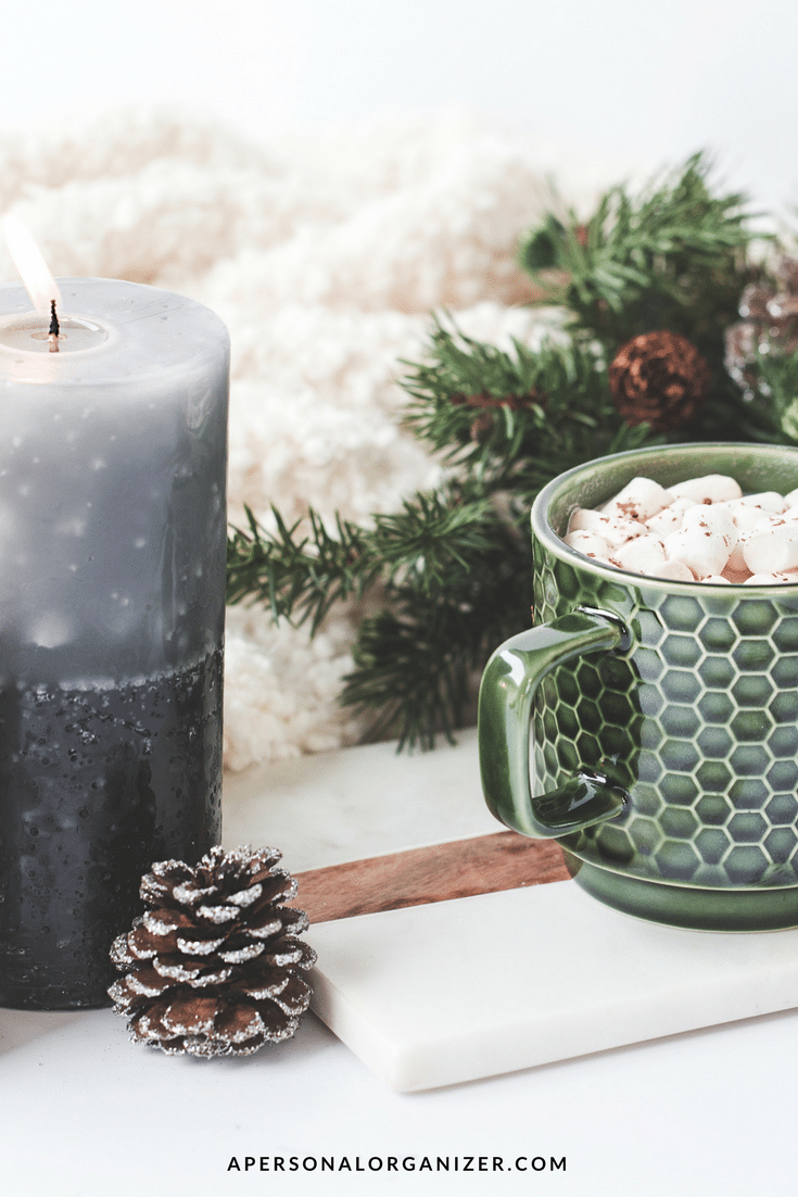A cozy scene features a green mug with a honeycomb pattern filled with marshmallow-topped hot chocolate. The mug sits on a white tray next to a pine cone and a lit gray candle, perfect for some holiday organizing in the background alongside evergreen branches and a fluffy white blanket.