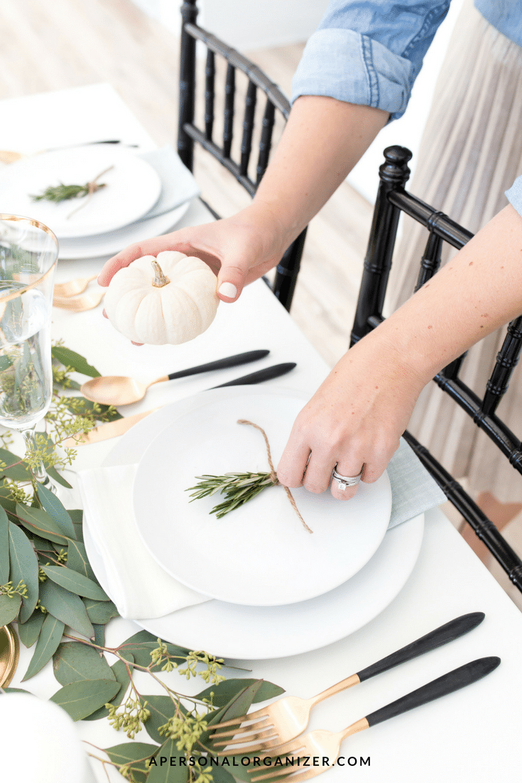 A person is carefully arranging a small pumpkin on a white plate set for a meal. The table, thoughtfully decorated with greenery and accented with gold and black utensils, reflects the spirit of planning for Thanksgiving dinner. Nearby, a glass stands ready, while the person wears a blue shirt and pleated skirt.